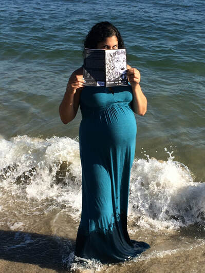 A pregnant Li Yun Alvarado wearing a long blue dress and holding her book, Words or Water, in front of her face, showing only her eyes.